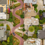 Aerial view of a residential neighborhood featuring a winding, brick-paved street lined with houses, greenery, and parked cars.