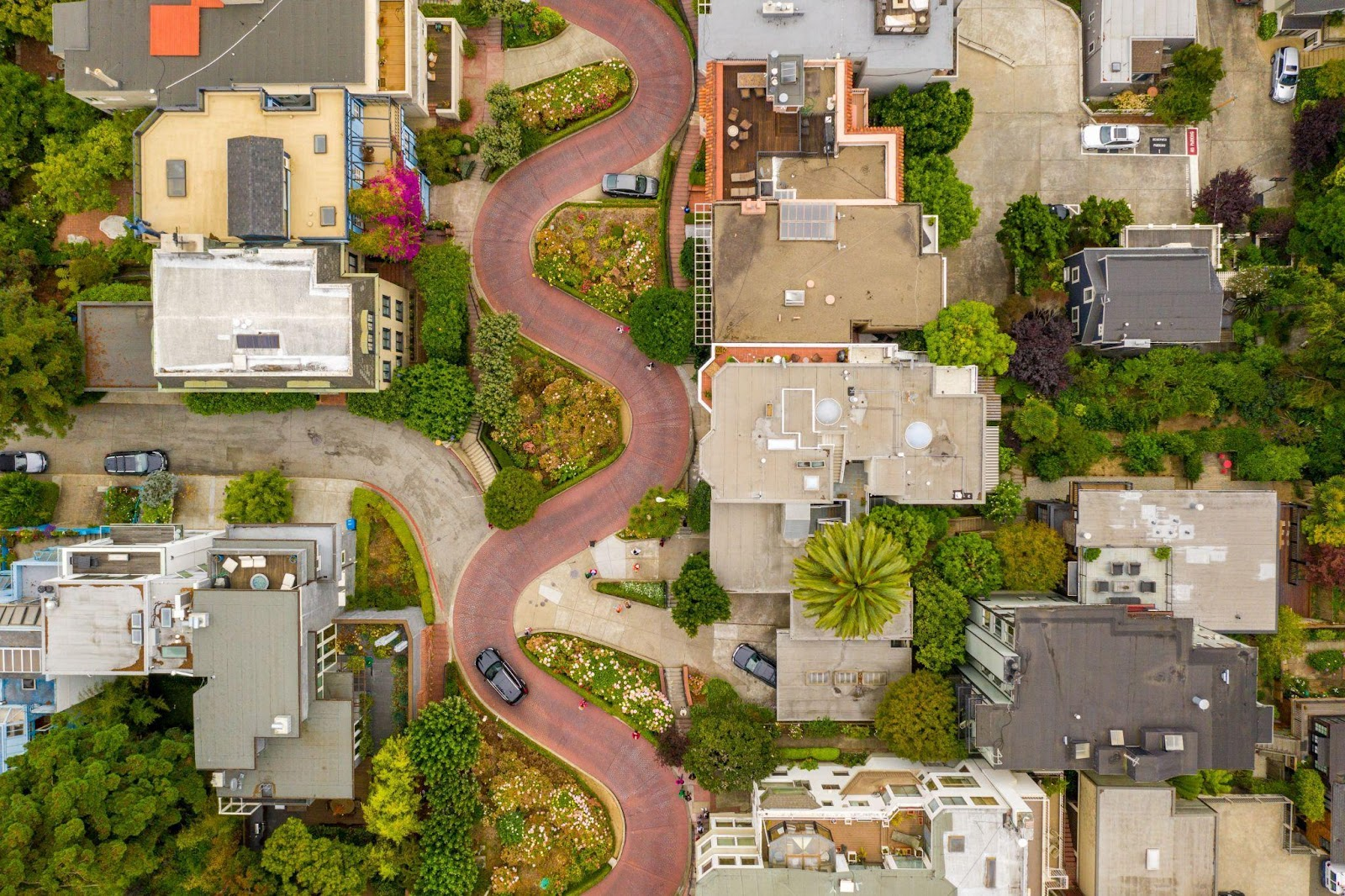Aerial view of a residential neighborhood featuring a winding, brick-paved street lined with houses, greenery, and parked cars.