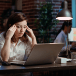 Woman in white shirt looking stressed while working on laptop at desk in office setting with exposed brick walls.