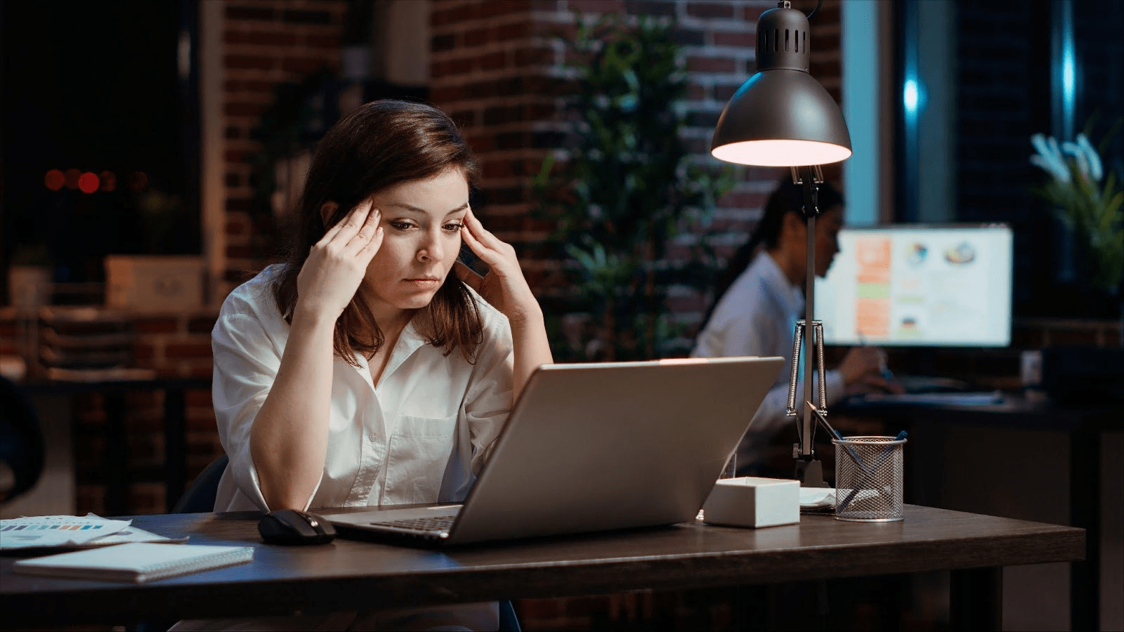 Woman in white shirt looking stressed while working on laptop at desk in office setting with exposed brick walls.