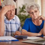 Senior couple looking concerned while reviewing financial documents with calculator at home table.