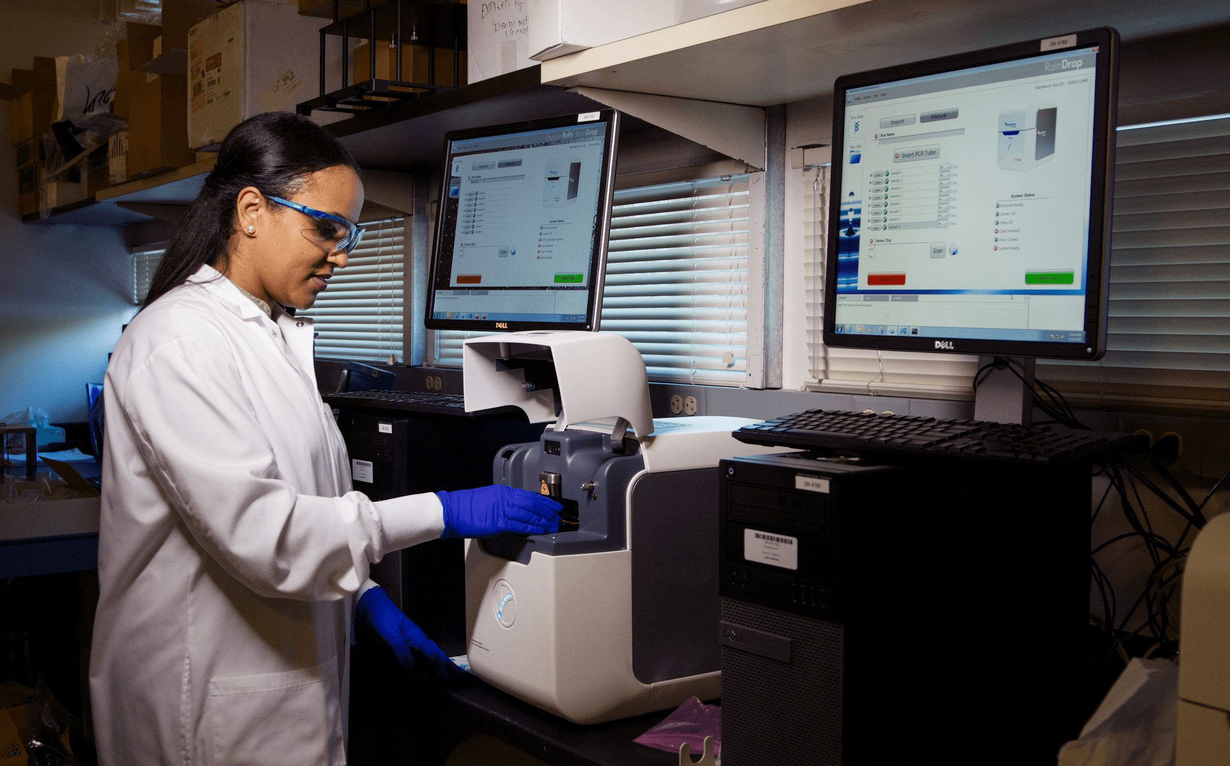 Female scientist in lab coat and safety gear operates scientific equipment. Two computer monitors display analysis software in the background.
