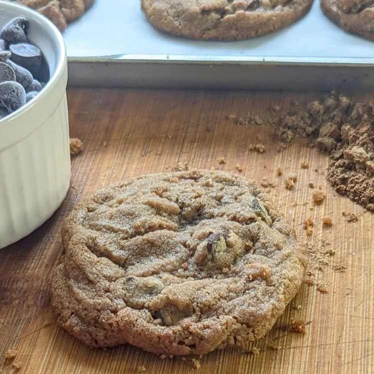 A gingerbread chocolate chip cookie on a wooden cutting board with gingerbread spice spilled on the board in the right center and a ramekin of chocolate chips to the left.