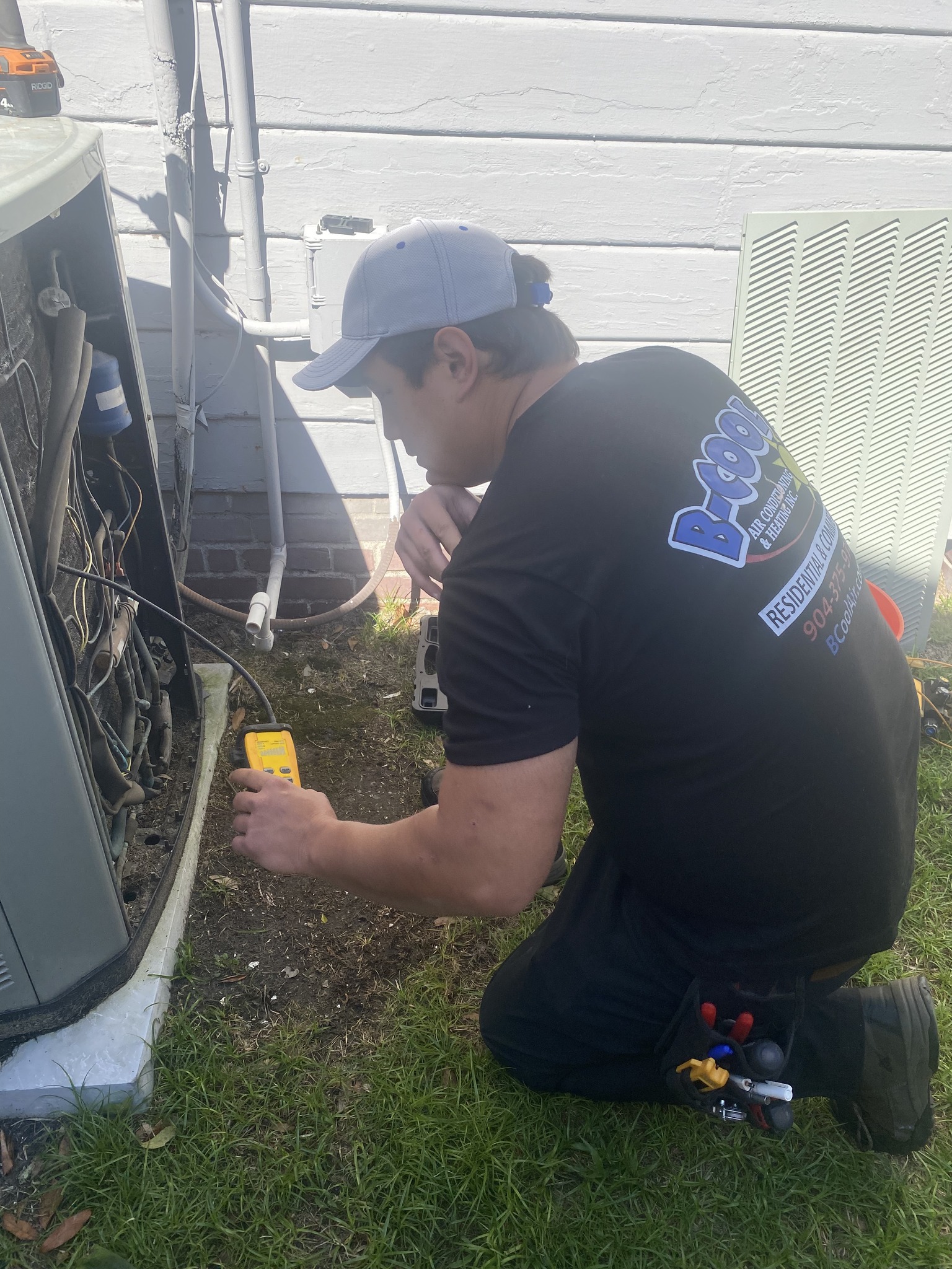 A technician in a black shirt and cap kneels on grass, inspecting an HVAC unit with a yellow meter tool.
