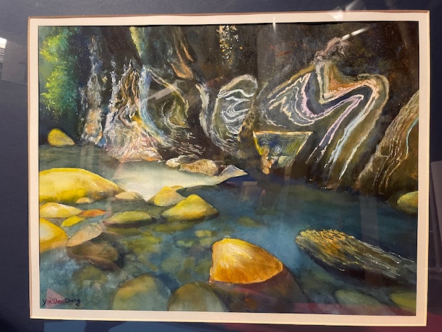 A watercolor painting by Yin Shen Chang featuring a shallow stream with smooth, sunlit yellow boulders in cool blue water. To the right, dark rocky canyon walls display distinct, swirling light-colored geological line patterns.
