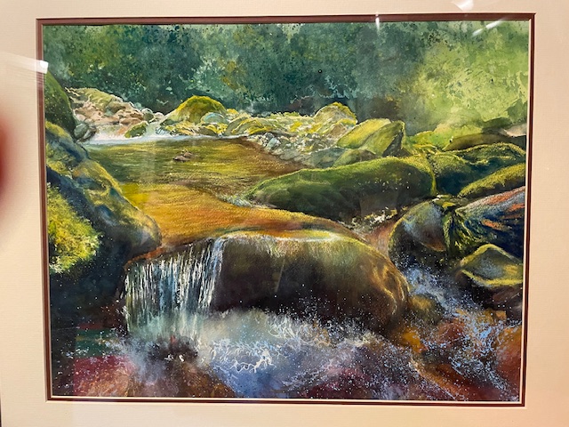 A watercolor painting depicting a small forest waterfall cascading over moss-covered dark boulders into a frothy splash pool. Warm sunlight filters through dense green foliage onto the calm stream upper pool and rocks.