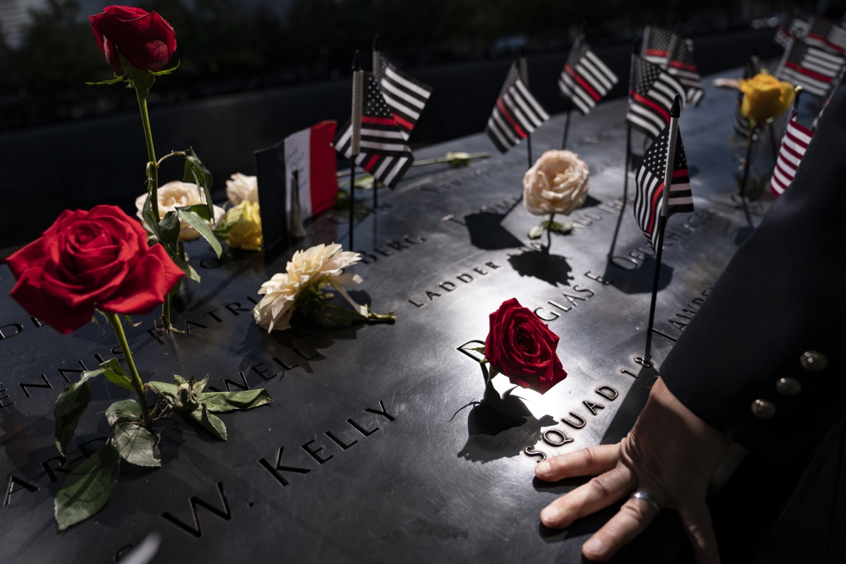 A person runs their hand over a dark grey headstone surrounded by roses and flags.