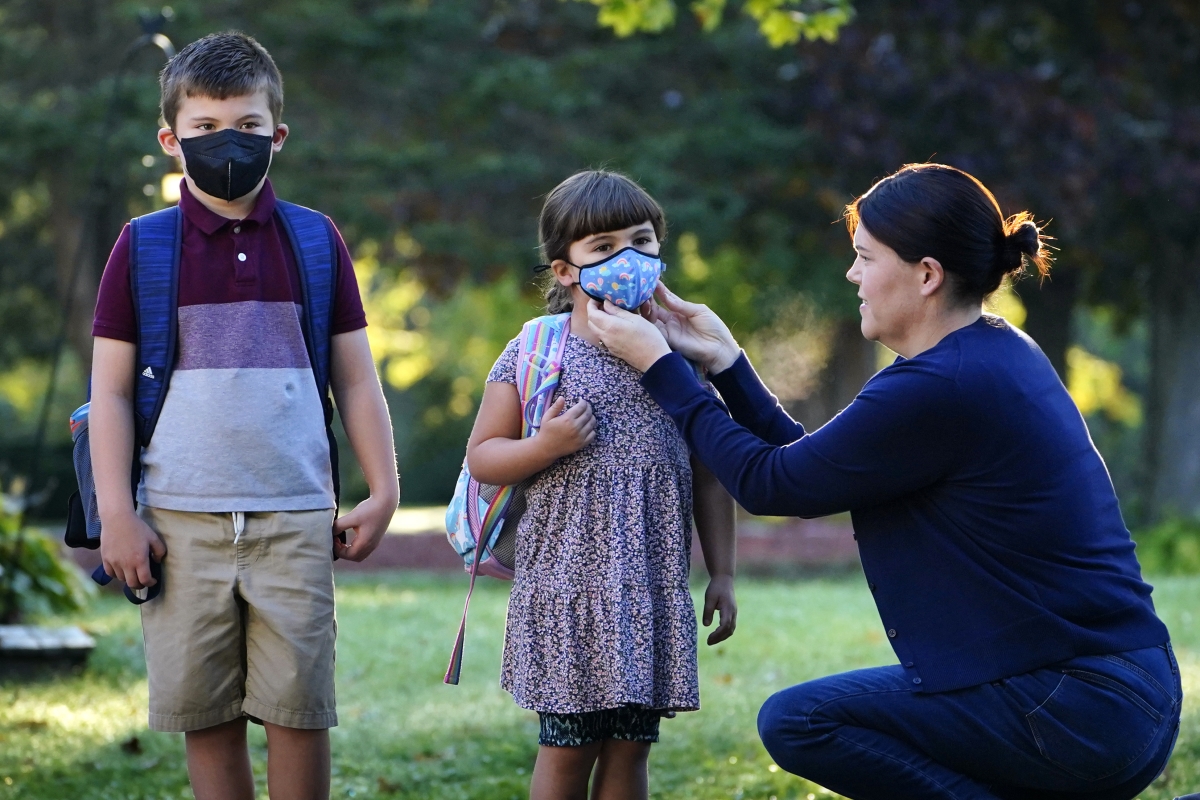 A young boy in a black face mask and a young girl in a patterned face mask stand with backpacks. Their mother, a woman wearing a blue shirt and pants with dark hair, adjusts the girl's face mask.