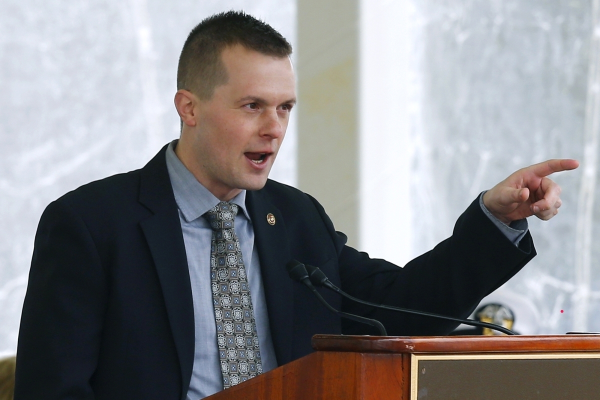 Jared Golden, a man with dark blond hair and dark eyes, wearing a navy blue blazer over a light blue button-down and a patterned tie, points from behind a wooden podium.