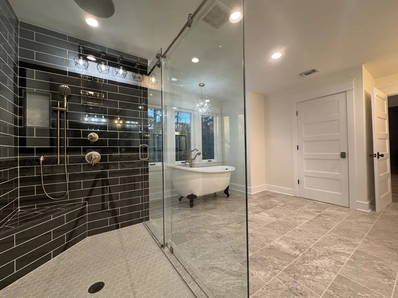 Walk-in shower with black subway tile, glass door, and freestanding tub beyond