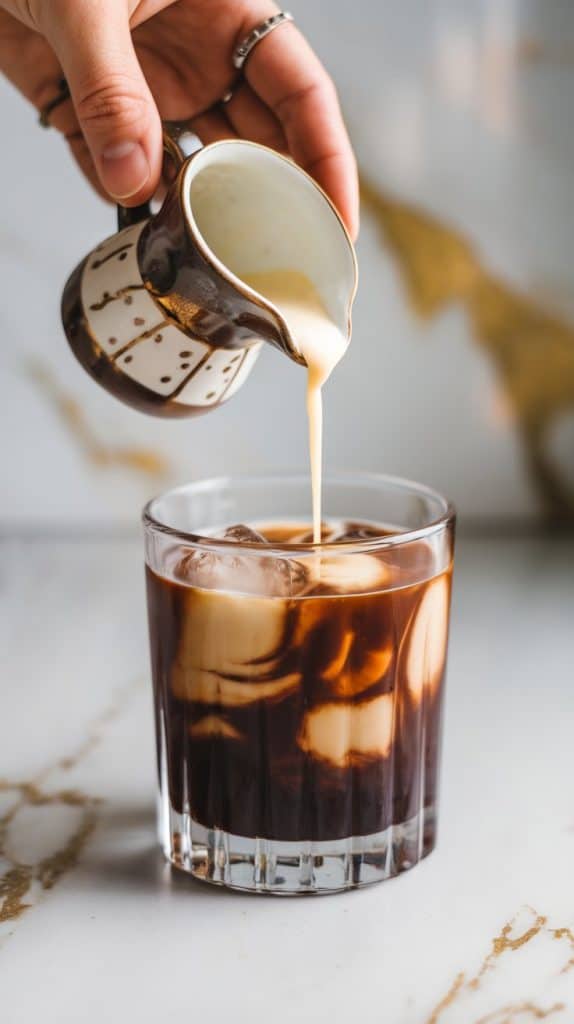 A hand pouring heavy cream from a small pitcher into a rocks glass filled with espresso, vodka, and coffee liqueur, creating soft swirls in the drink.