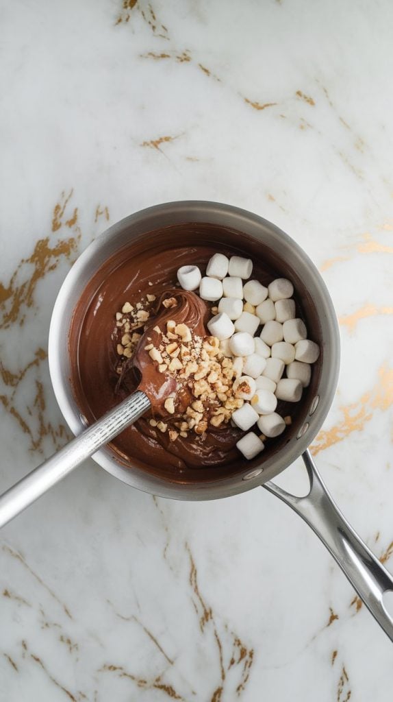 A saucepan off the heat with smooth chocolate fudge mixture being folded together with mini marshmallows and chopped nuts.