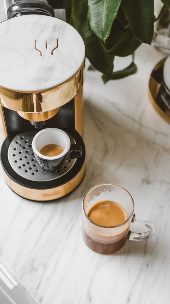 A small espresso cup being poured into a tall glass mug, with a coffee maker, raspberry syrup bottle, and chocolate syrup nearby on a white marble counter with hints of gold.