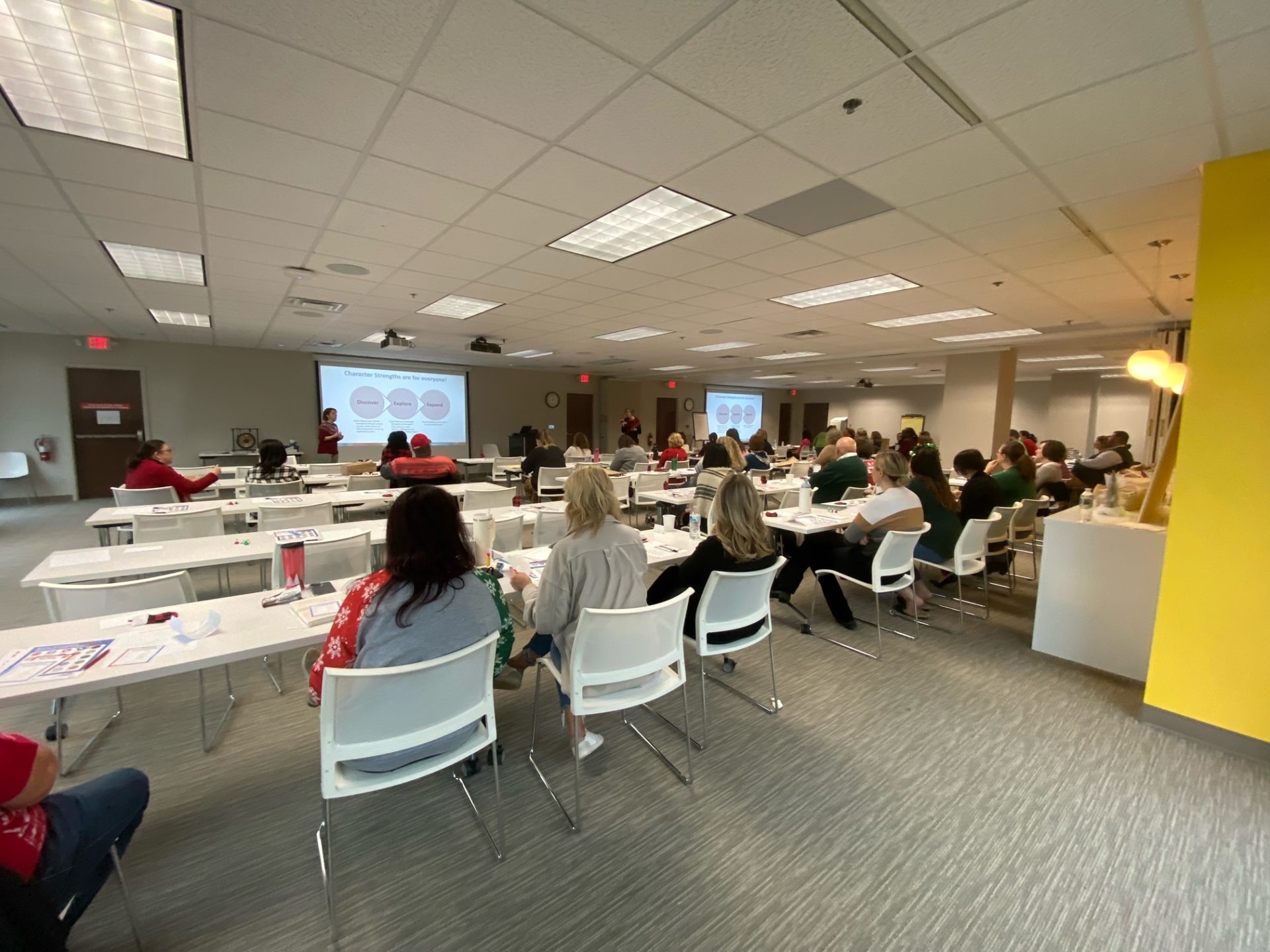 Photo of a group of people sitting in white chairs in a classroom looking at a person giving a PowerPoint presentation in the front of the room