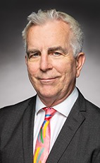 Professional headshot of a man in a suit, smiling, with gray hair and a colorful tie.