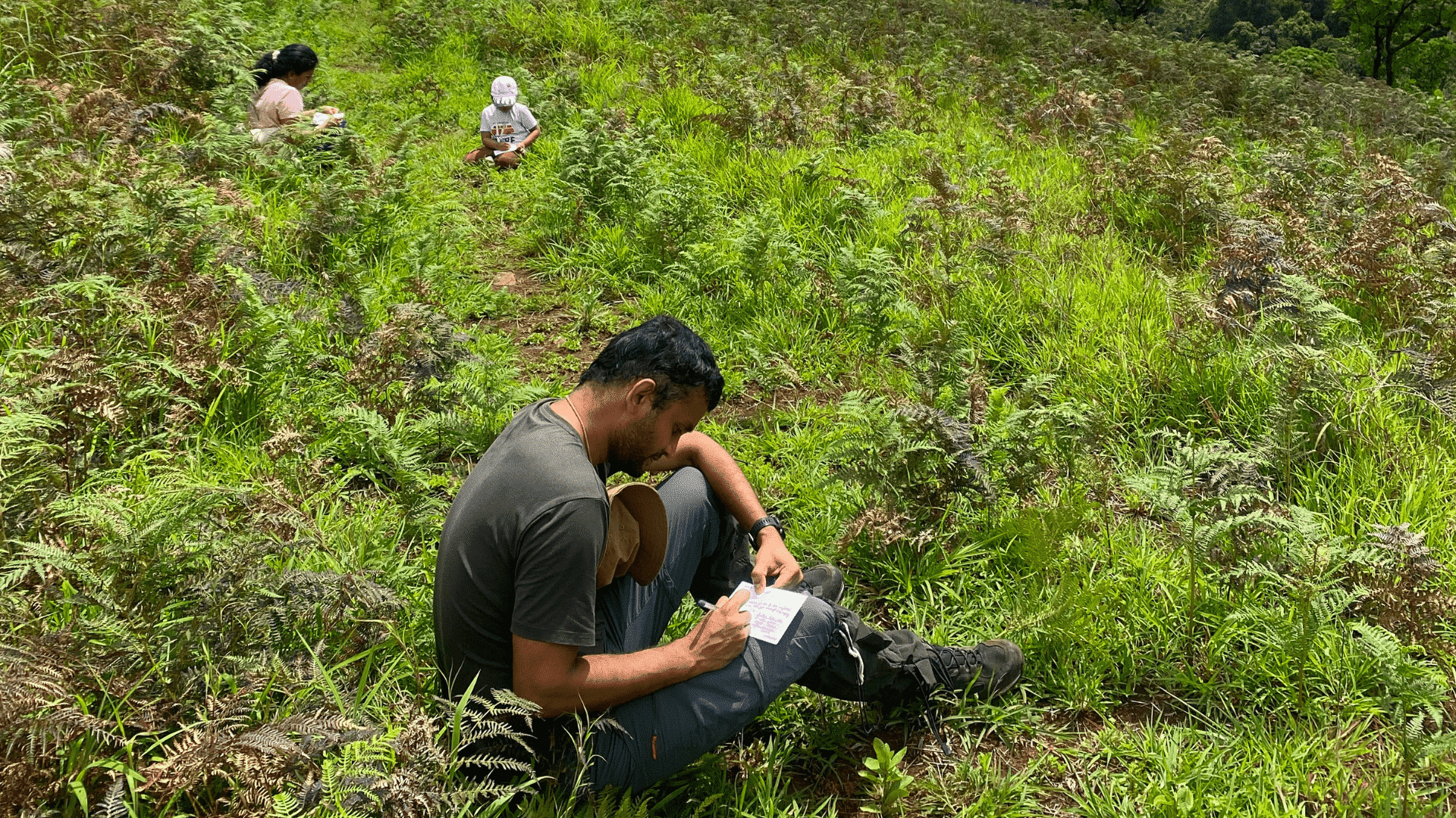 A man sits among lush green ferns and grass, writing notes during a field activity, while a woman and child observe the landscape in the background.