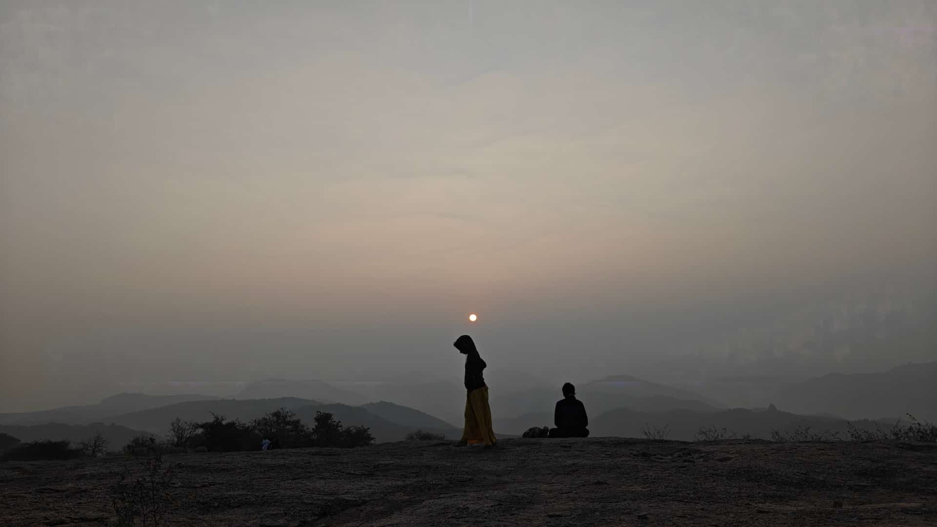Two people silhouetted on a rocky hilltop, one standing and one seated, with hazy mountain ranges and a low sun behind them.