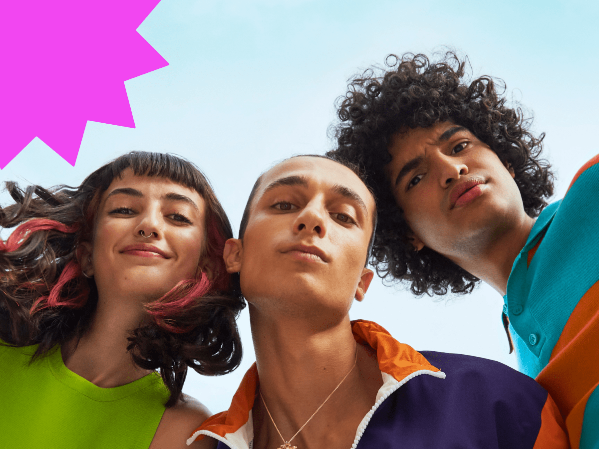 Three confident Gen Z friends looking down at the camera against a bright sky, dressed in bold, colorful outfits, representing diversity, self-expression, and connection.