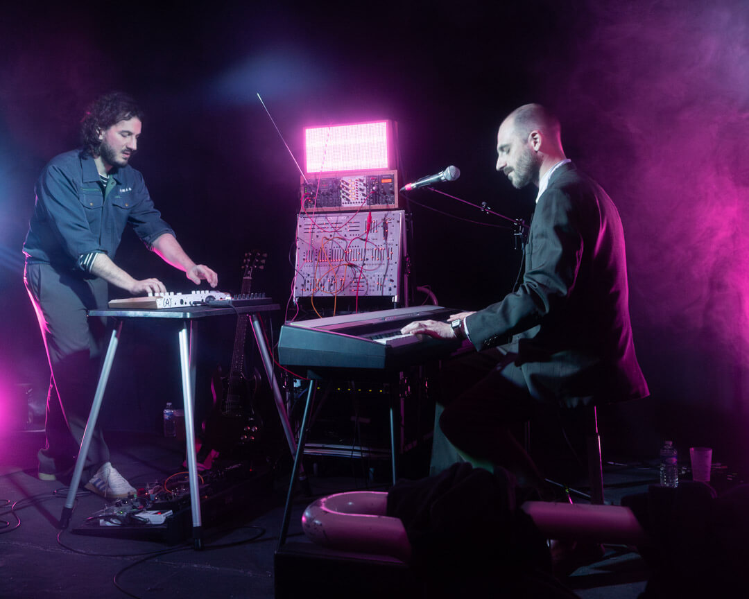 men duet playing a concert; the one on the left using a controler and the one on the right playing on a keyboard, pink smoke and light flares in the frame