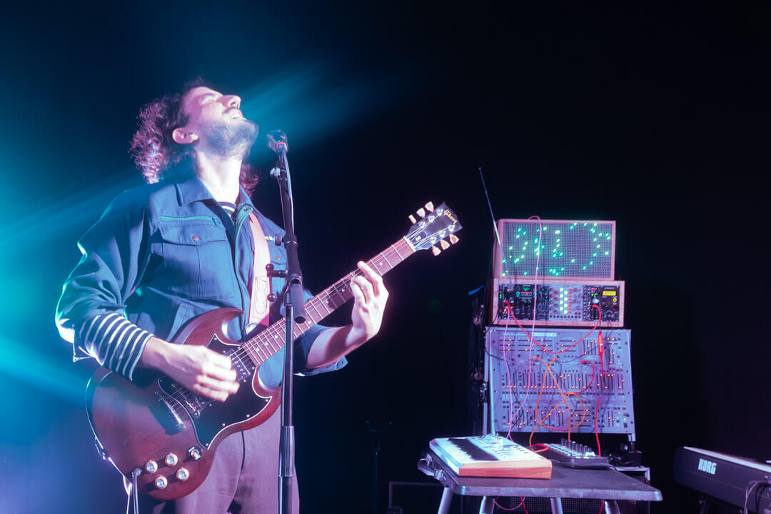 artistic concert photo of a man playing an electronic guitar surrounded by electronic y2k equipment and with a keyboard