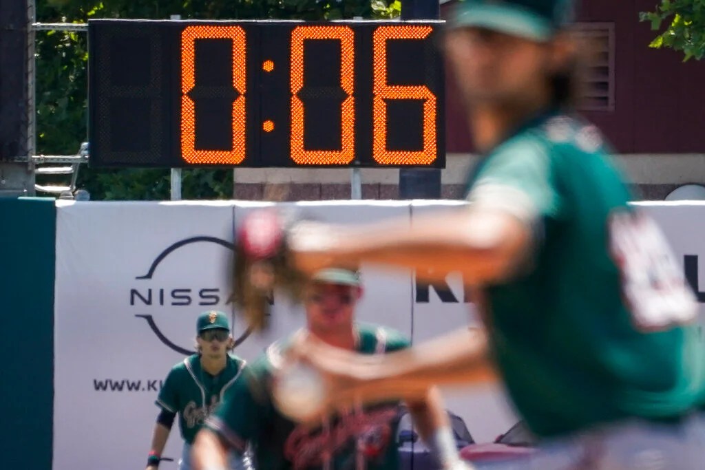 Reloj digital del estadio marcando 0:06, con un pelotero de uniforme verde en posición de bateo desenfocado en primer plano