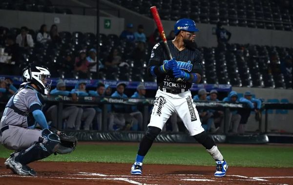 Bateador en uniforme azul y blanco con casco azul en posición de bateo, receptor agachado de fondo, durante un juego de beisbol en estadio