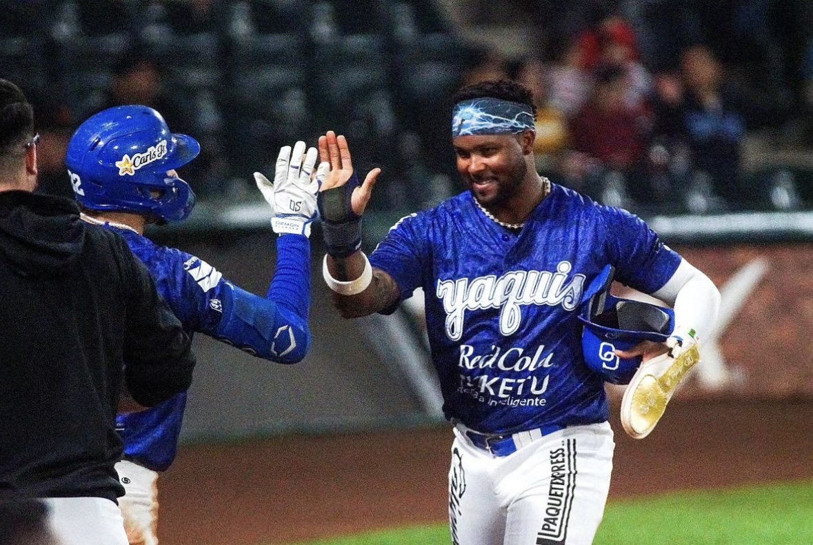 Pelotero de Yaquis en uniforme azul celebra con compañero tras anotar carrera en el juego