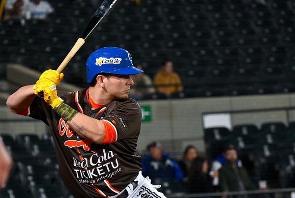 Bateador con casco azul y uniforme negro con franjas rojas en posición de bateo, sosteniendo el bate en el hombro durante un juego de beisbol en estadio