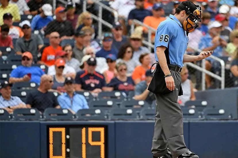 Árbitro en uniforme azul y gris revisa su reloj mientras el cronómetro del estadio marca 0:19 durante un juego de beisbol