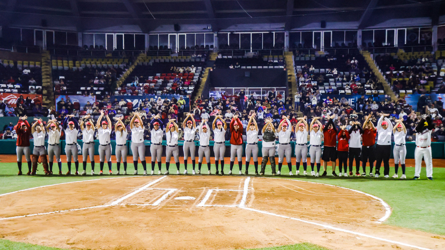Equipo femenil de beisbol alineado en el terreno durante presentación de roster, con jugadoras en uniforme blanco y algunas en rojo, ante estadio lleno