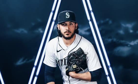 Lanzador con uniforme blanco y azul marino de los Marineros sostiene su guante de beisbol en posición de lanzamiento, con fondo de cielo nublado y luces de neón azules