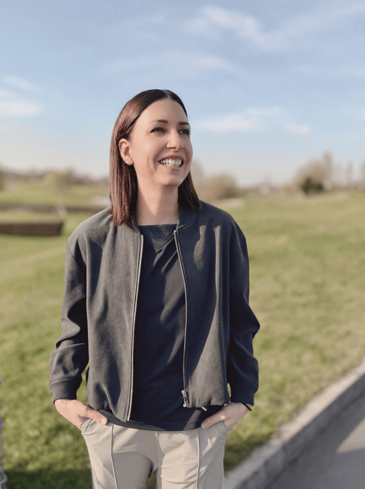 Portrait of a smiling woman on a golf course. She is wearing a dark grey bomber jacket over a dark T-shirt and beige pleated trousers. The background is a vast green lawn under a blue sky, presented in a soft, cinematic lifestyle look.