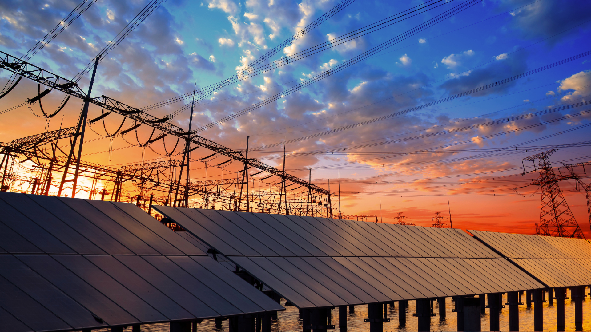 Solar panels in front of a high-voltage power grid at sunset.