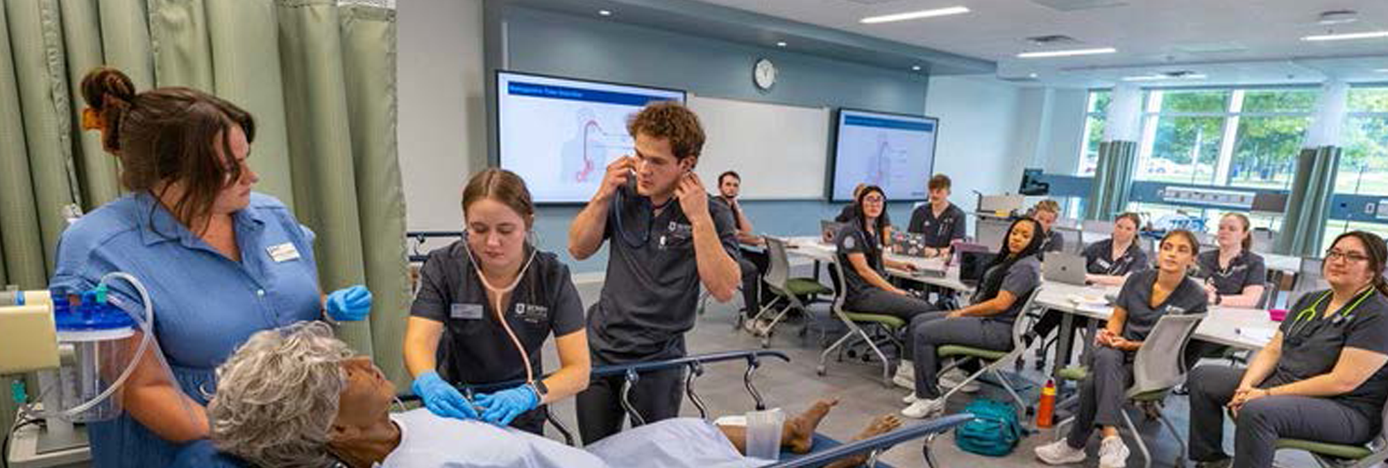 A medical training session in a classroom setting, featuring a nursing team attending to a simulated patient. The scene shows multiple students actively engaged in learning with a focus on practical skills, while others observe. The environment includes medical equipment and screens displaying educational content.