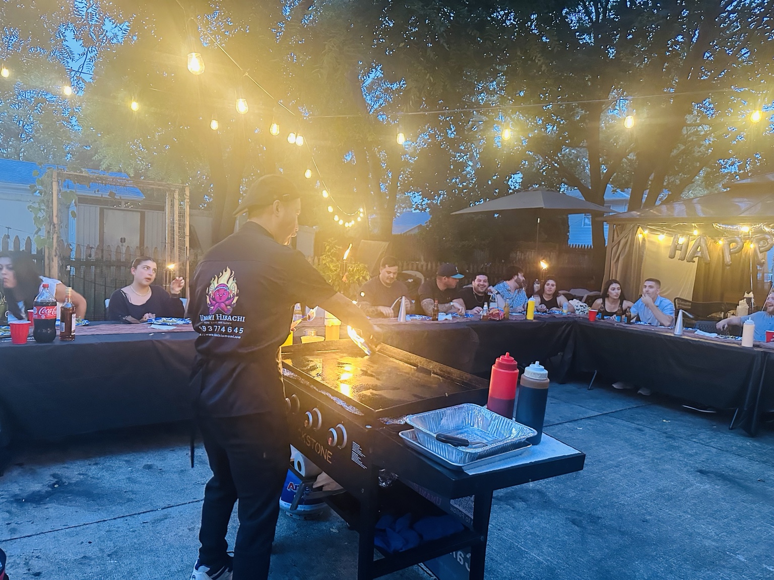 Outdoor hibachi chef and guests dining around the grill under string lights