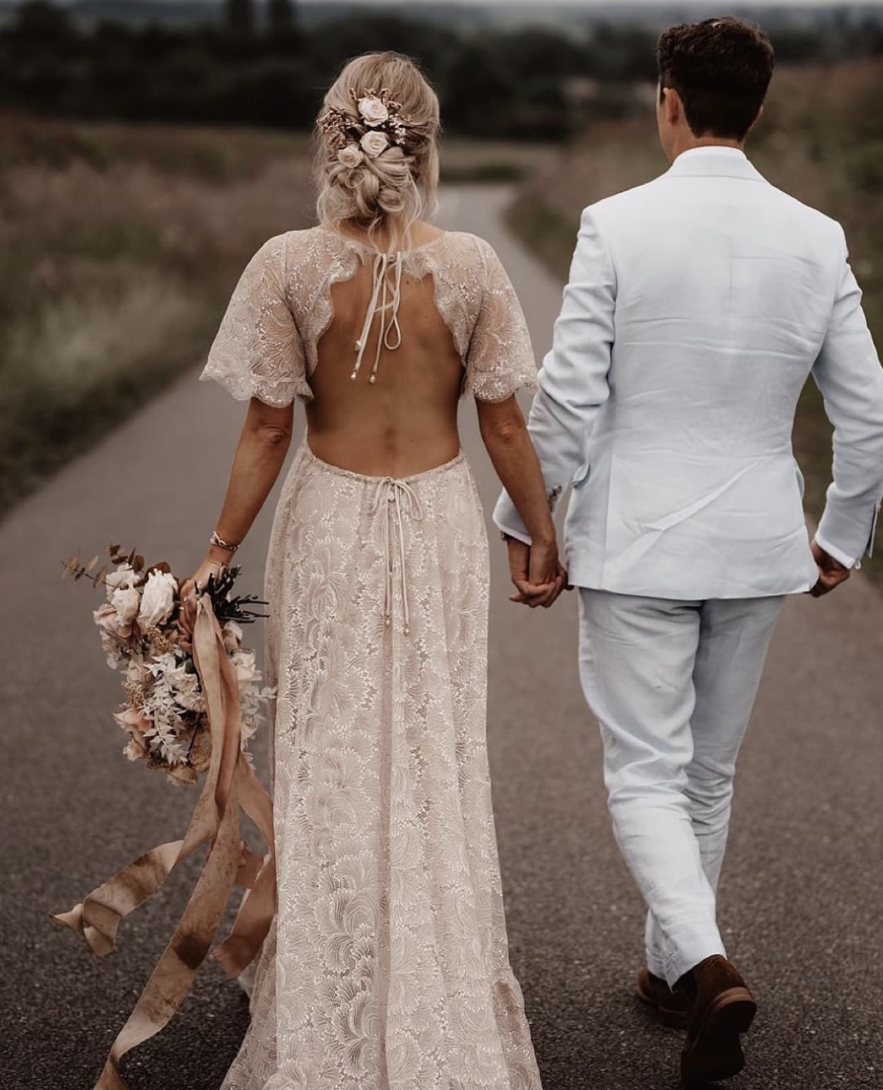 woman in lace wedding dress with bouquet holding hands with man in white suit walking down road