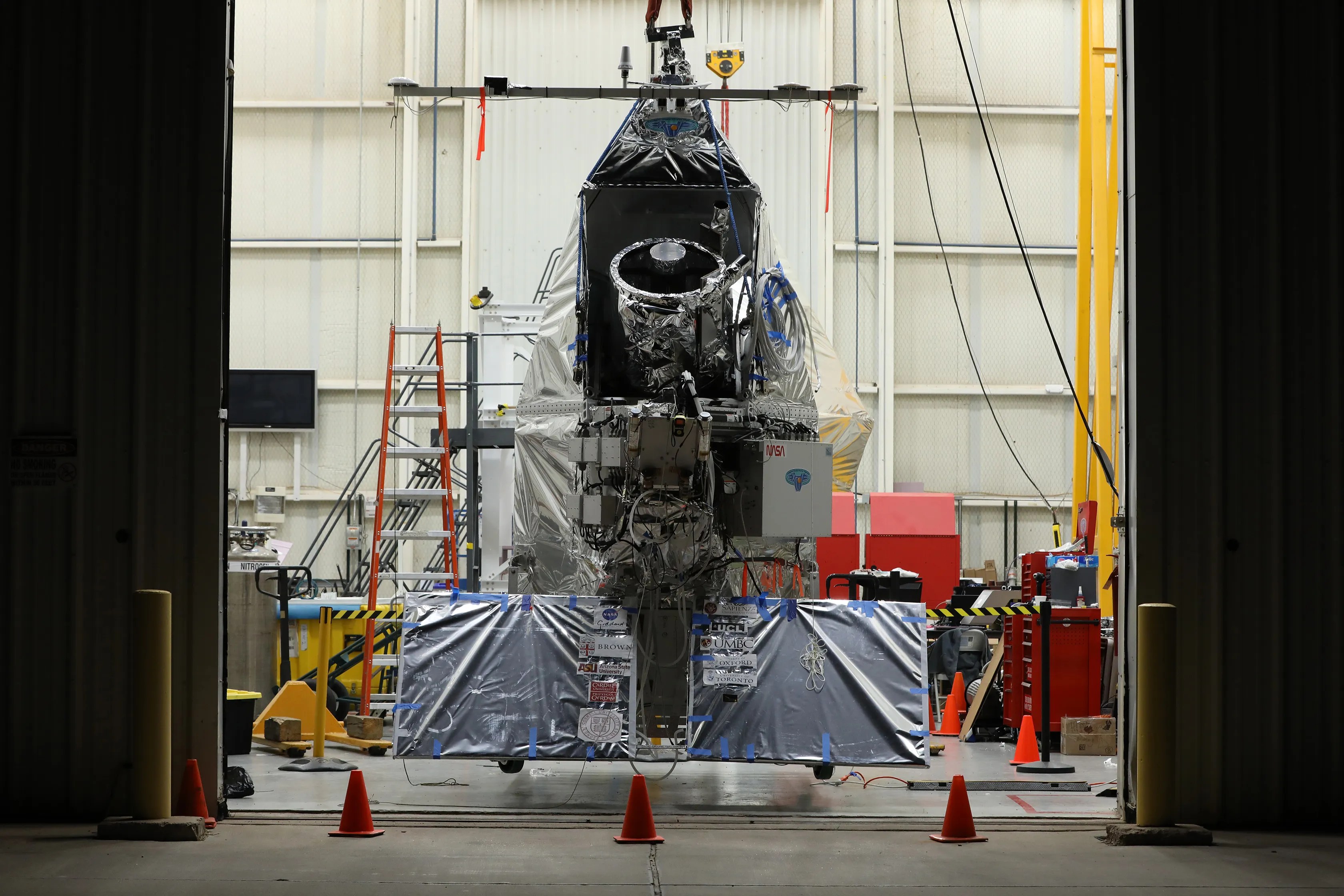 A satellite wrapped in reflective material is suspended in a large industrial facility, with orange cones and equipment visible in the background.