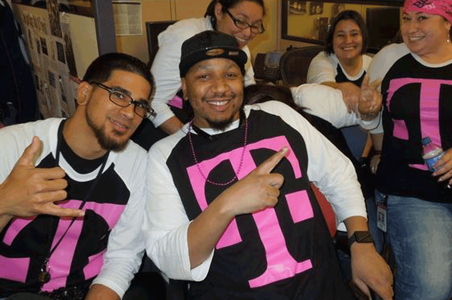 A joyful group of T-Mobile care agents is shown, playfully seated in front of the camera in a break room. 1 of the 5 people shown is making a shaka hand-sign, one is making a peace sign, and the individual in the foreground is pointing and smiling to the person making a peace sign. All individuals shown are wearing long-sleeved raglan shirts with giant pink "T" T-Mobile logos on each.