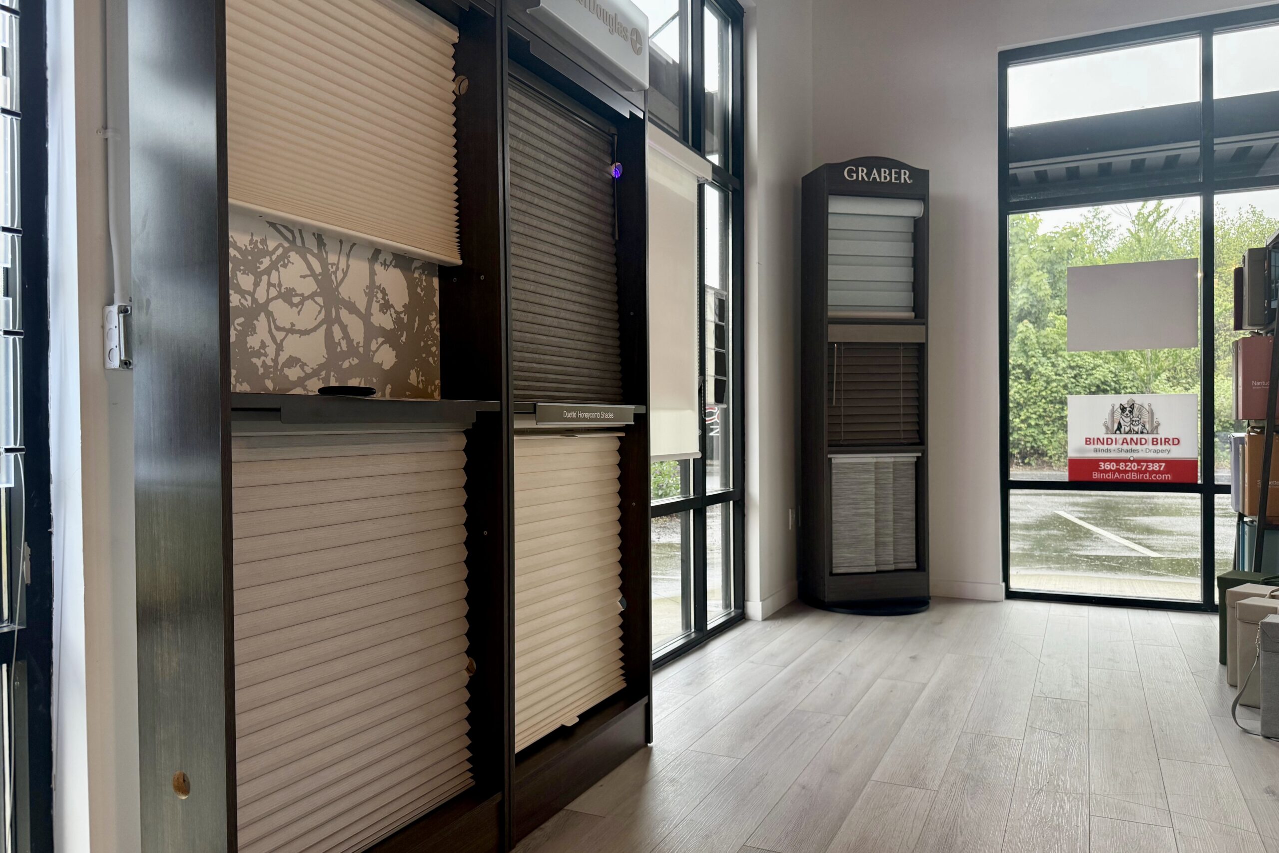 Showroom displaying various window blinds and shades on tall dark wood displays, with large windows to the right and a 'Graber' sample unit nearby.