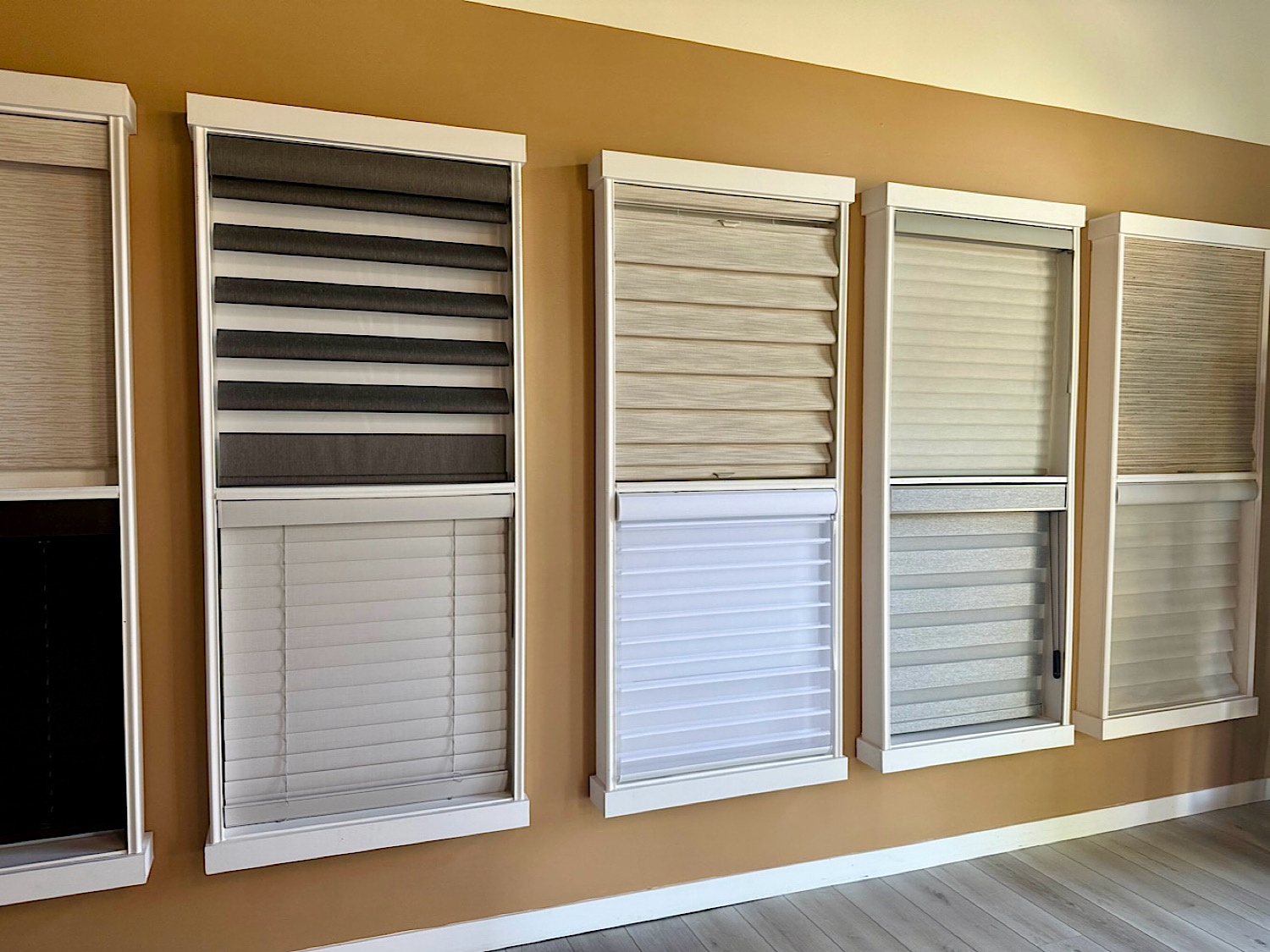 Row of window blind samples mounted on a showroom wall, featuring horizontal slat blinds in white, beige, and gray tones.
