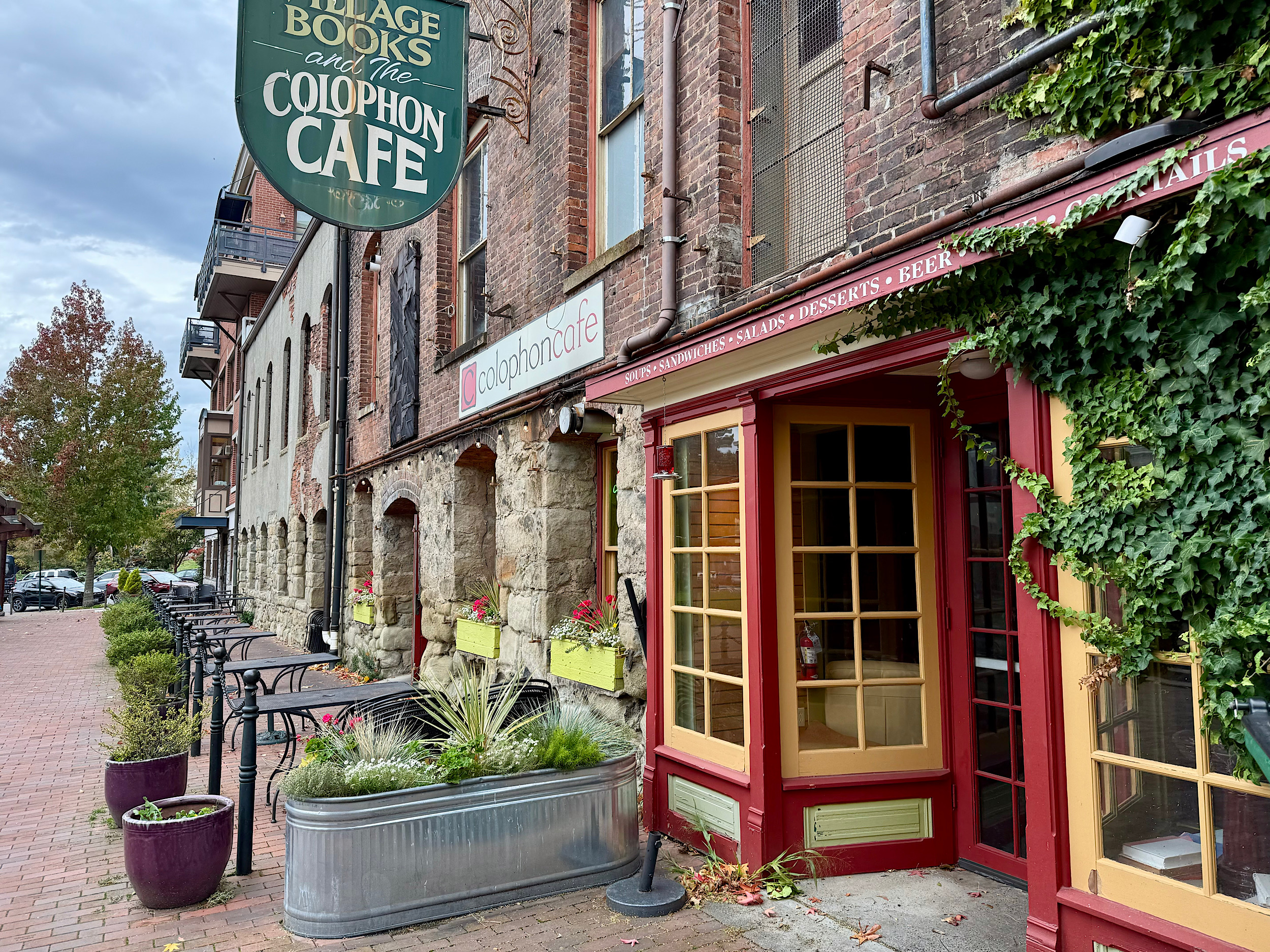 Brick storefront with ivy, red/yellow window frames, and outdoor seating along a brick sidewalk; sign reads Colophon Café.