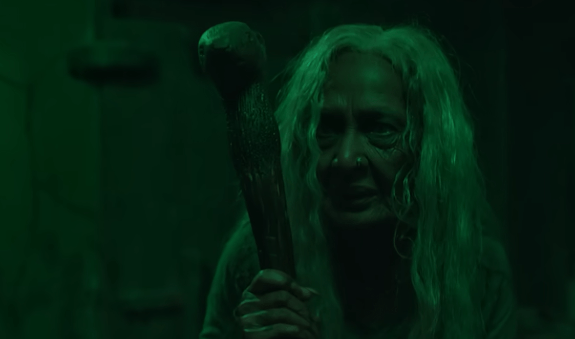 Muthassi elder character in green-lit folk-horror scene