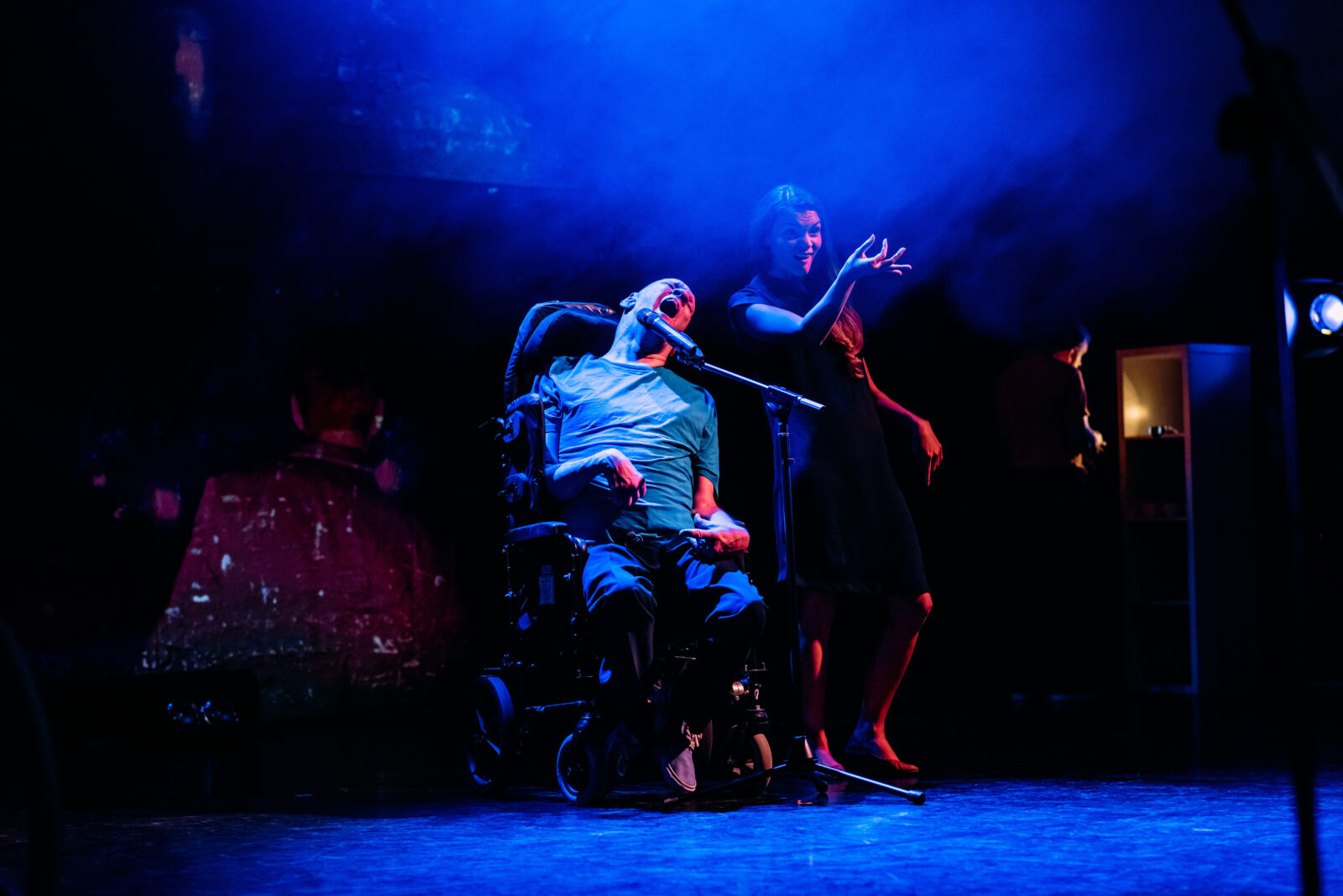 A middle aged man sits in his wheelchair and talks into a microphone. The stage is lit a dark blue. Next to the man is a young woman signing.