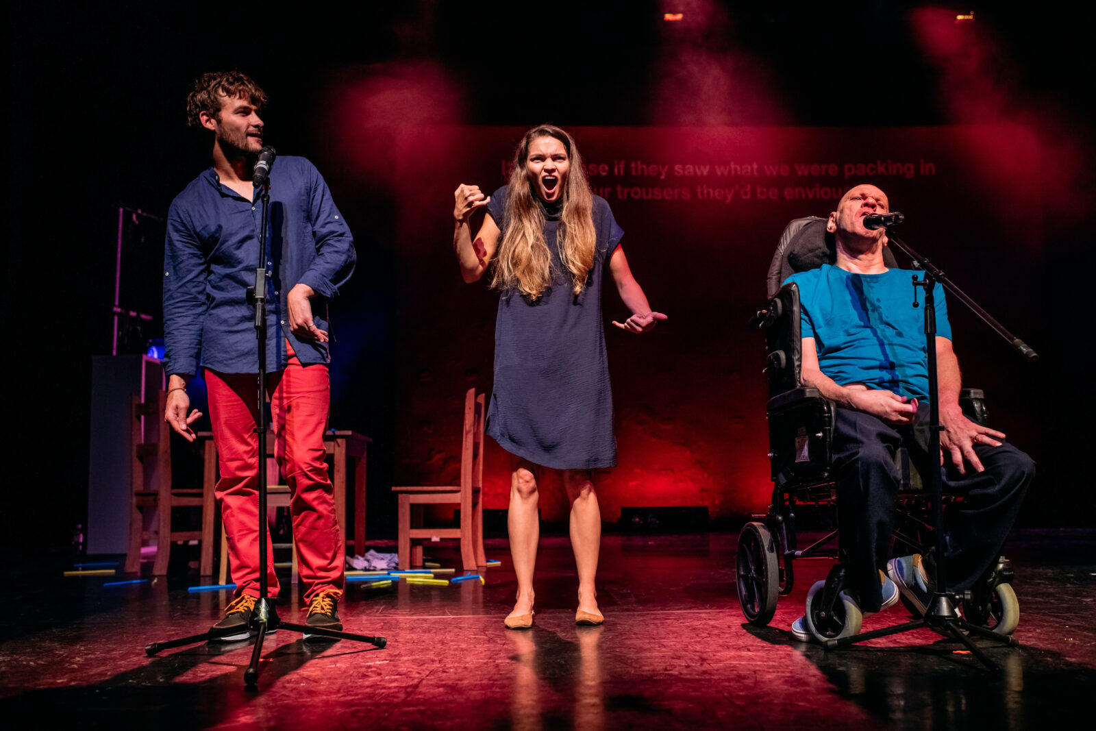 Two men line up in front of microphones at the front of the stage that has been lit red. There is a woman in between them, she has long blonde hair and is wearing a blue dress. She appears to be in the middle of signing and her mouth is open wide in a shocked expression.