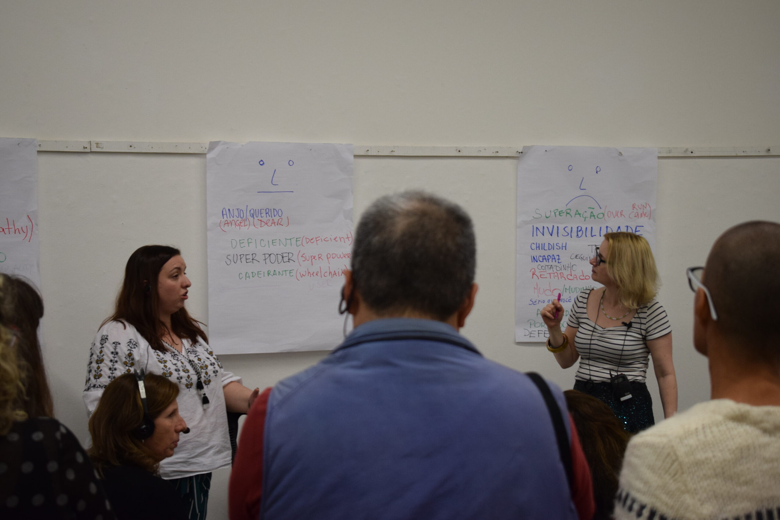 Two young women stand beside large pieces of paper stuck to the wall. They are holding pens as if they have just been writing on the pages. We see the backs of people's heads, watching them.