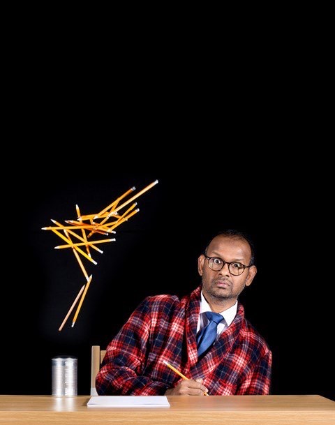 Ramesh is a middle aged Sri Lankan man wearing glasses, a red chechered suit jacket and blue tie. He sits at a desk but his body is angled diagonally at 45 degress. He holds a pencil and appears to be writing on a sheet of paper. A pot of pencils next to him seems to have erupted, spoting pencils into the air.