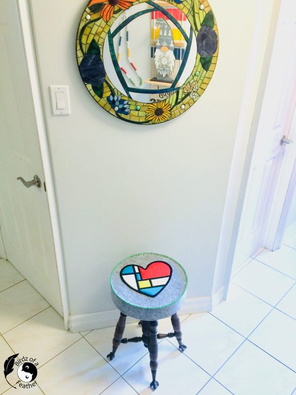 felt applique Mondrian heart stool cover on vintage wooden piano stool show below a mosaic mirror. Felt colours are grey, red, white blue, yellow and black felt with blanket stitched edge