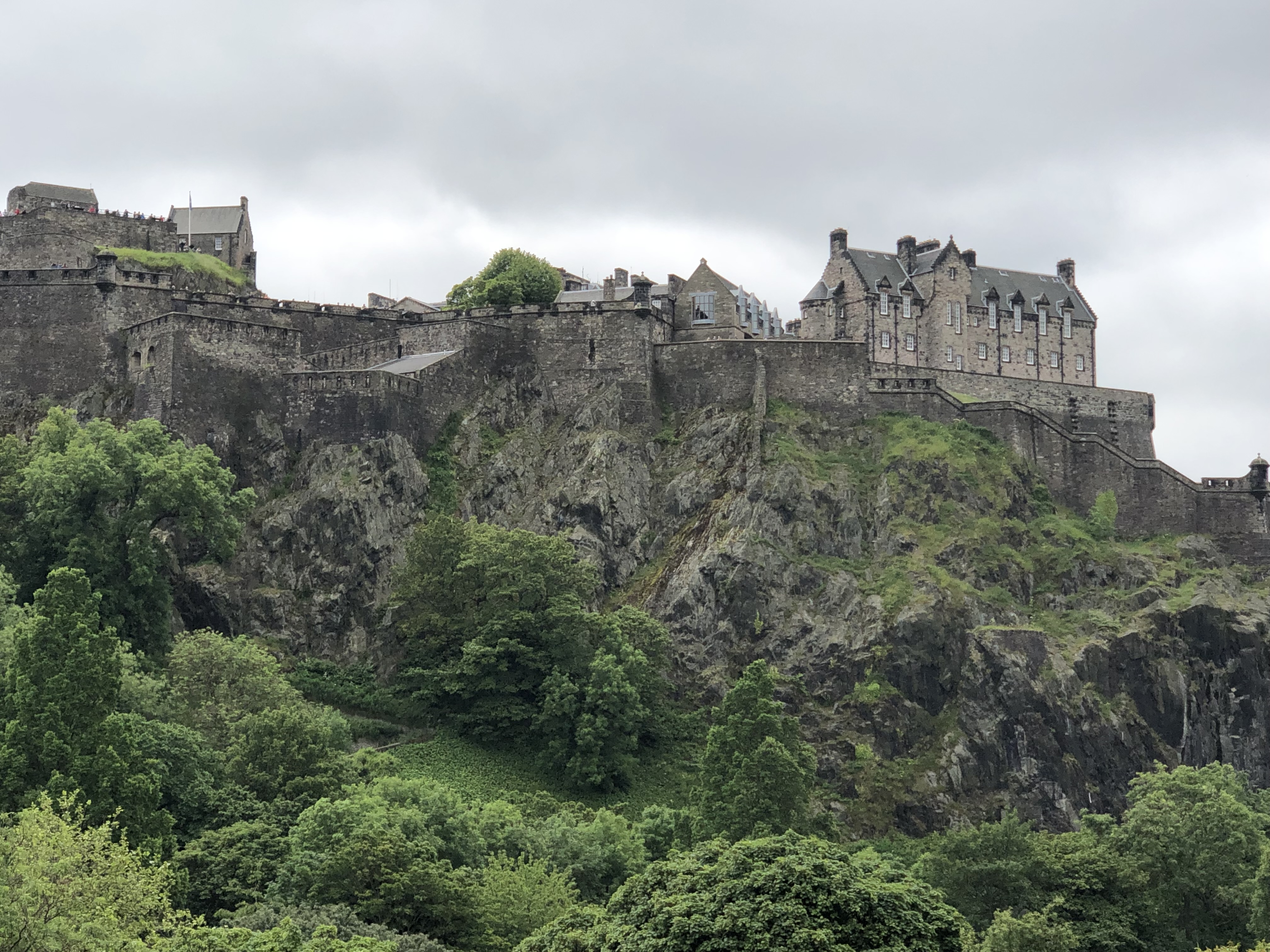 Photo of a castle up on a cliff. Cliff is covered in green bushes and trees. Castle is gray.