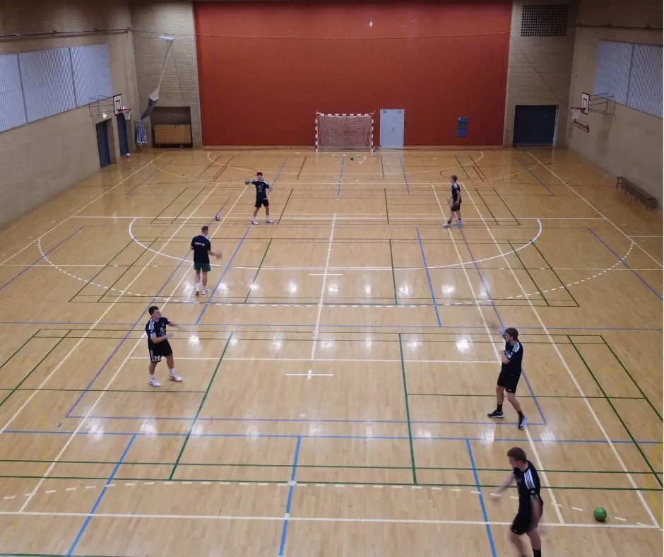Indendørs sportshal med trægulv, hvor flere håndboldspillere træner på tværs af banen i Hal 2 i Bjerringbro Idræts- & Kulturcenter.