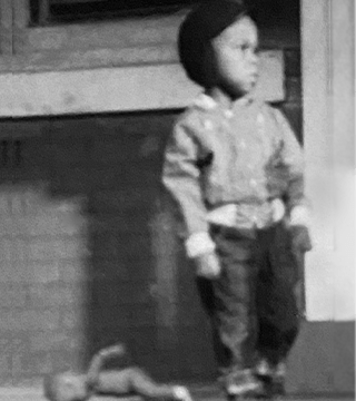 A black-and-white childhood photograph of Mark Ruffin standing on a porch, looking outward, with his first doll resting on the ground nearby.